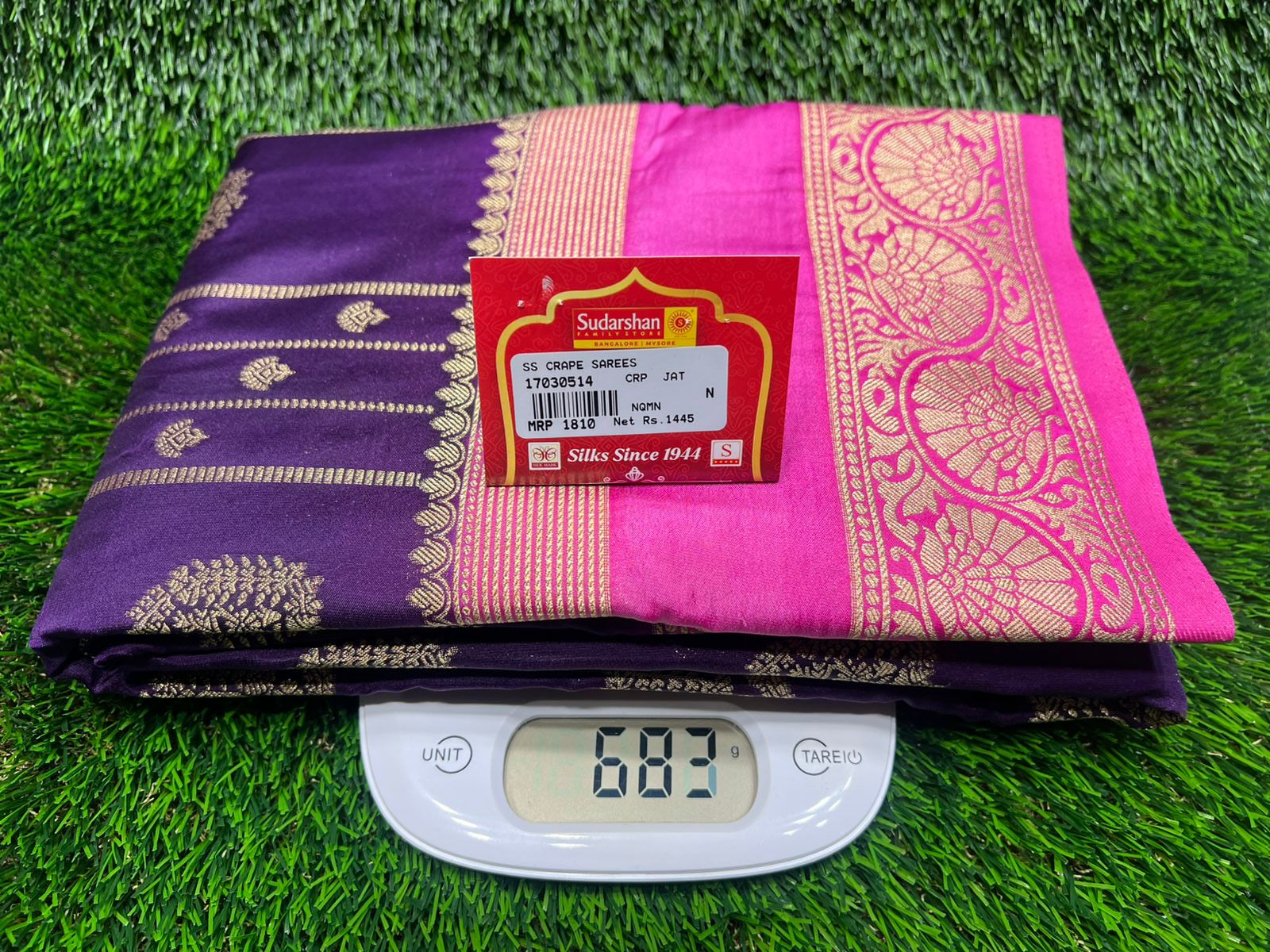 Purple and pink saree with a digital weight scale on a green grass background