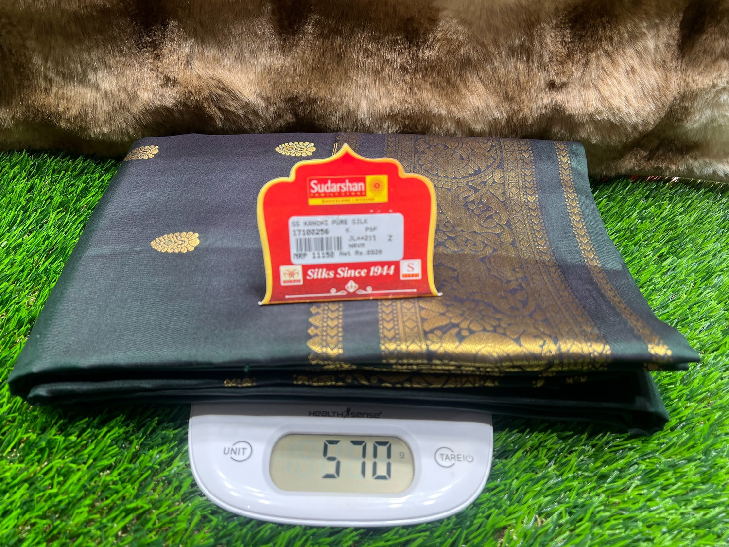 Black silk fabric with a Sudarshan label on a digital scale, placed on green grass.