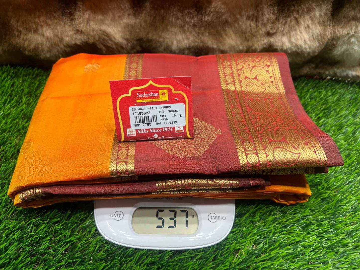 Folded red and orange saree with a label on a digital scale against a grassy background