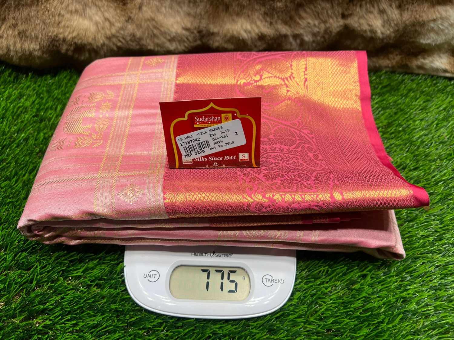 Pink saree with a brand label on a digital scale against a grass background