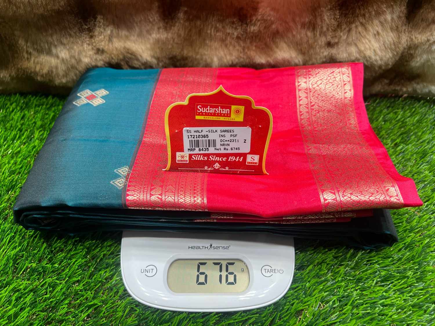 Two folded sarees, one blue and one pink with a label, on a digital scale displaying weight.