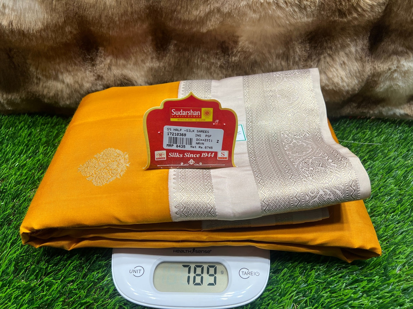 Folded yellow and white saree with a brand label on a digital scale against a grassy background
