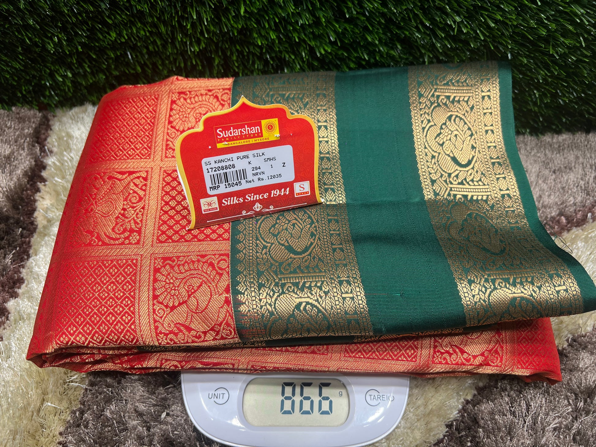 Red and green saree with a brand label on a digital scale against a grassy background
