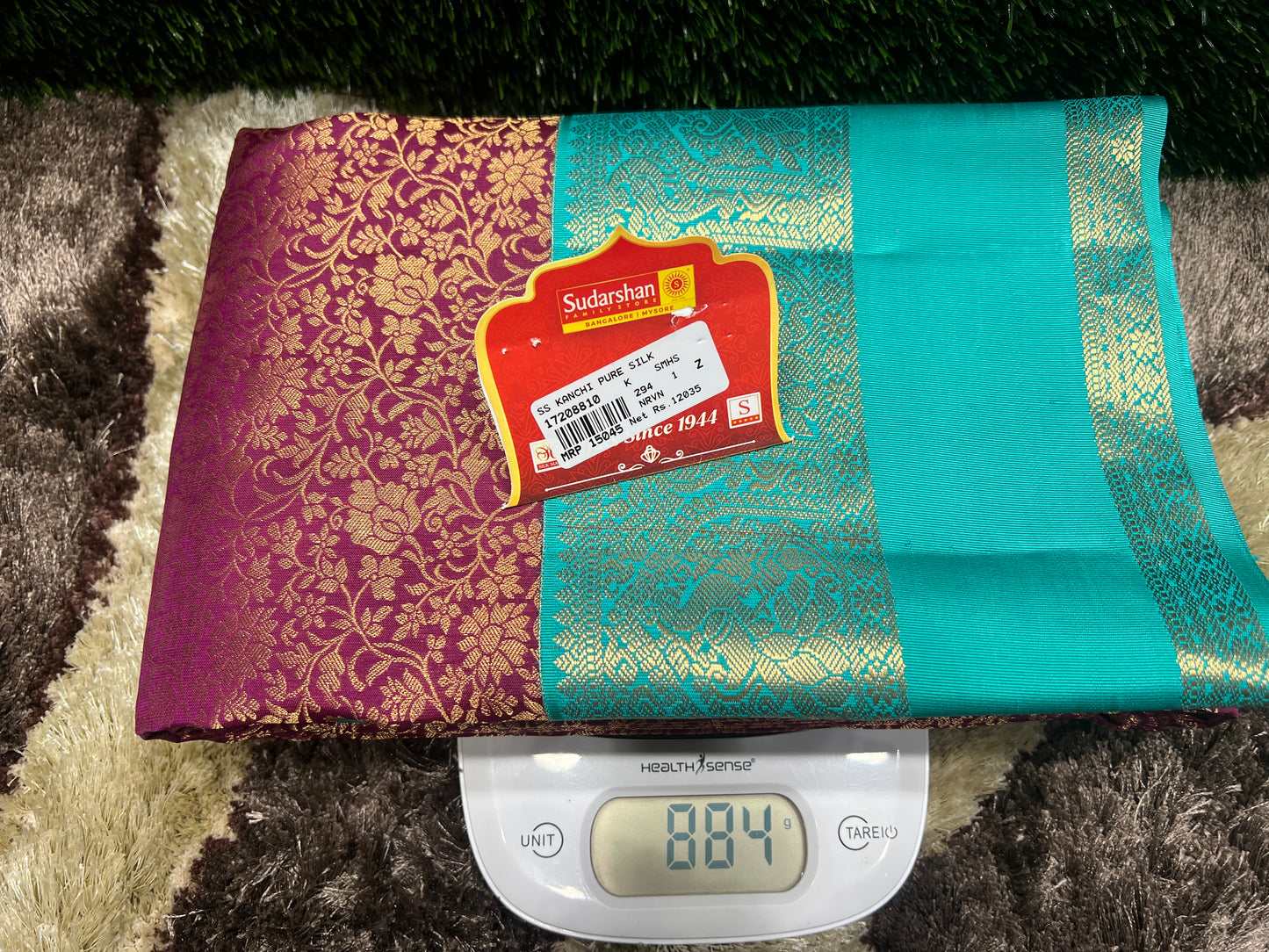 Two sarees, one purple and one teal with gold patterns, on a digital scale showing weight.