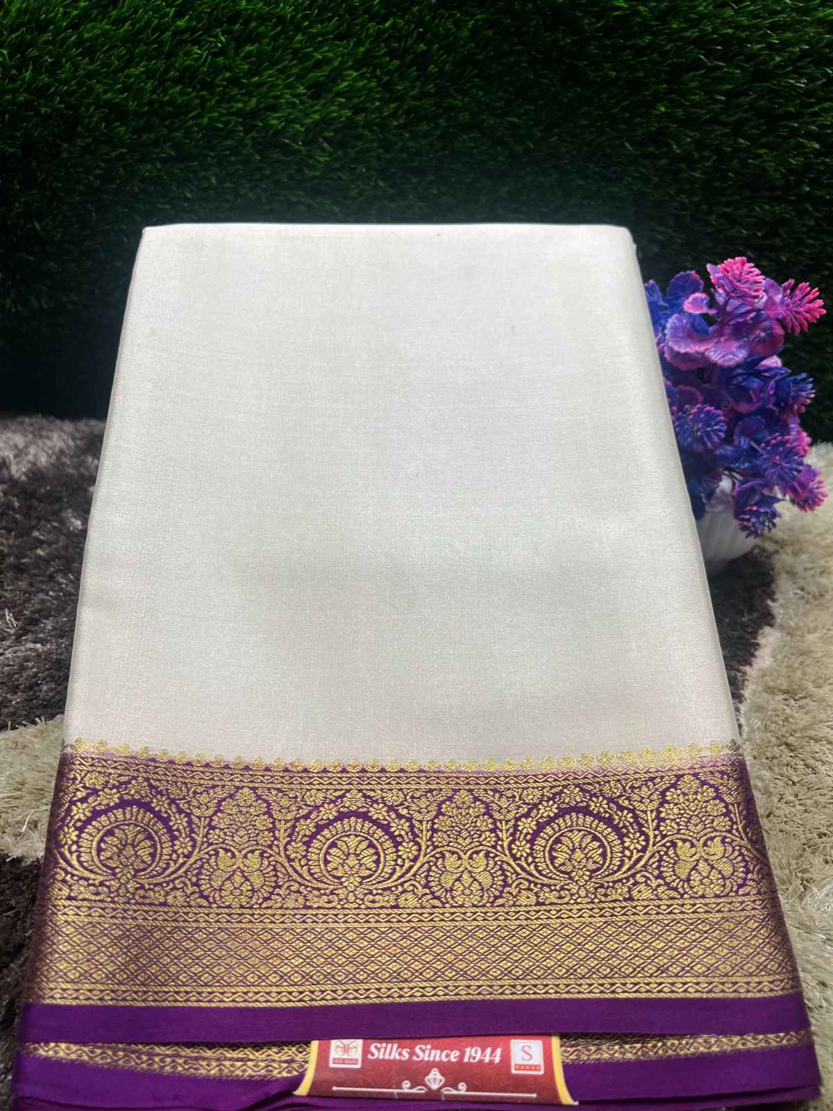 White saree with a purple and gold border on a textured surface with flowers in the background