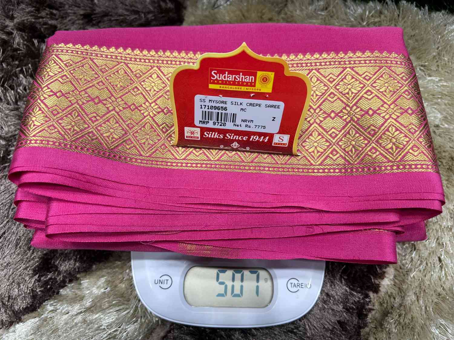 Pink fabric with gold patterns on a scale, featuring Sudarshan brand label.