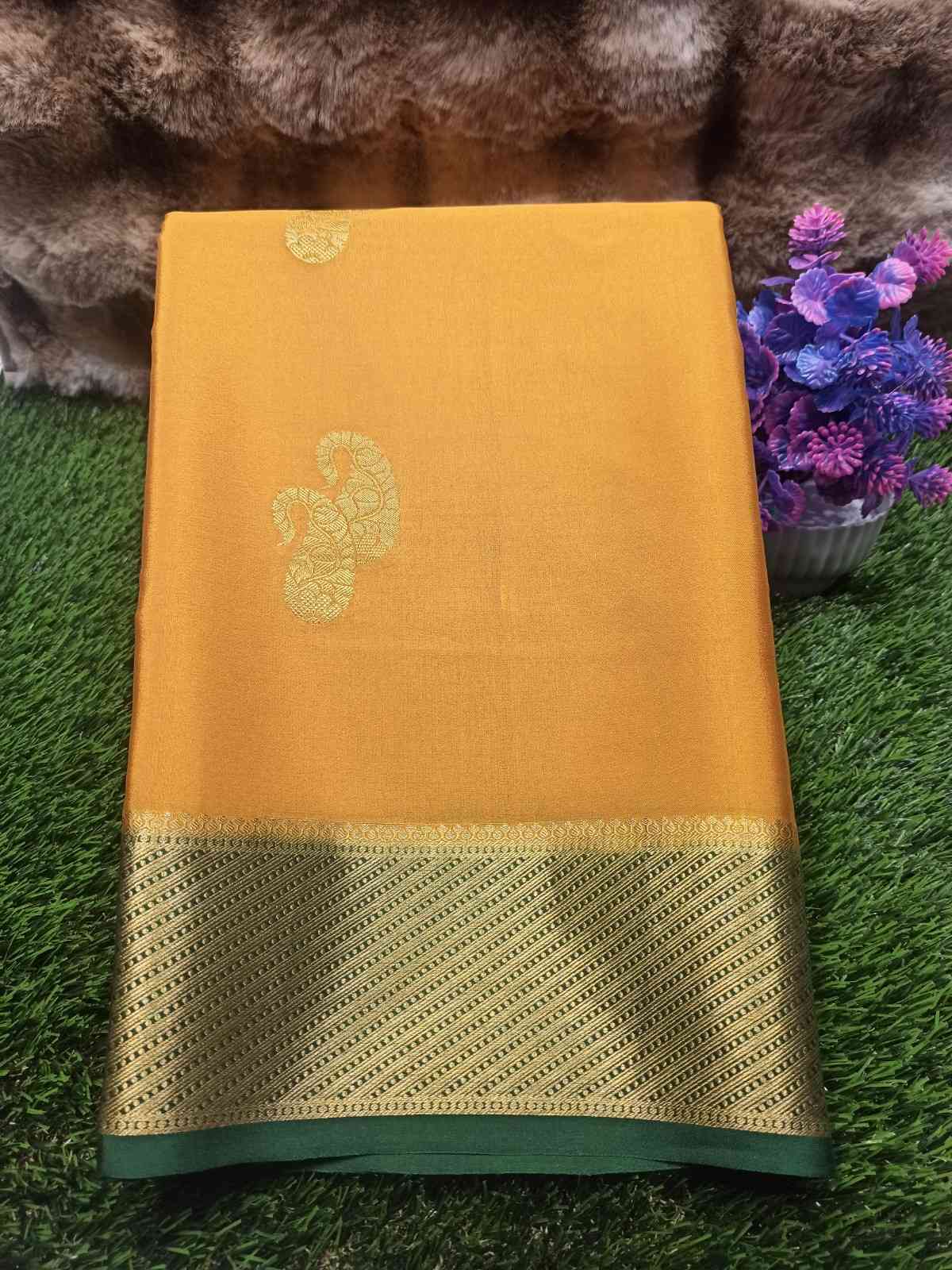 Yellow saree with gold border on a green surface with flowers in the background
