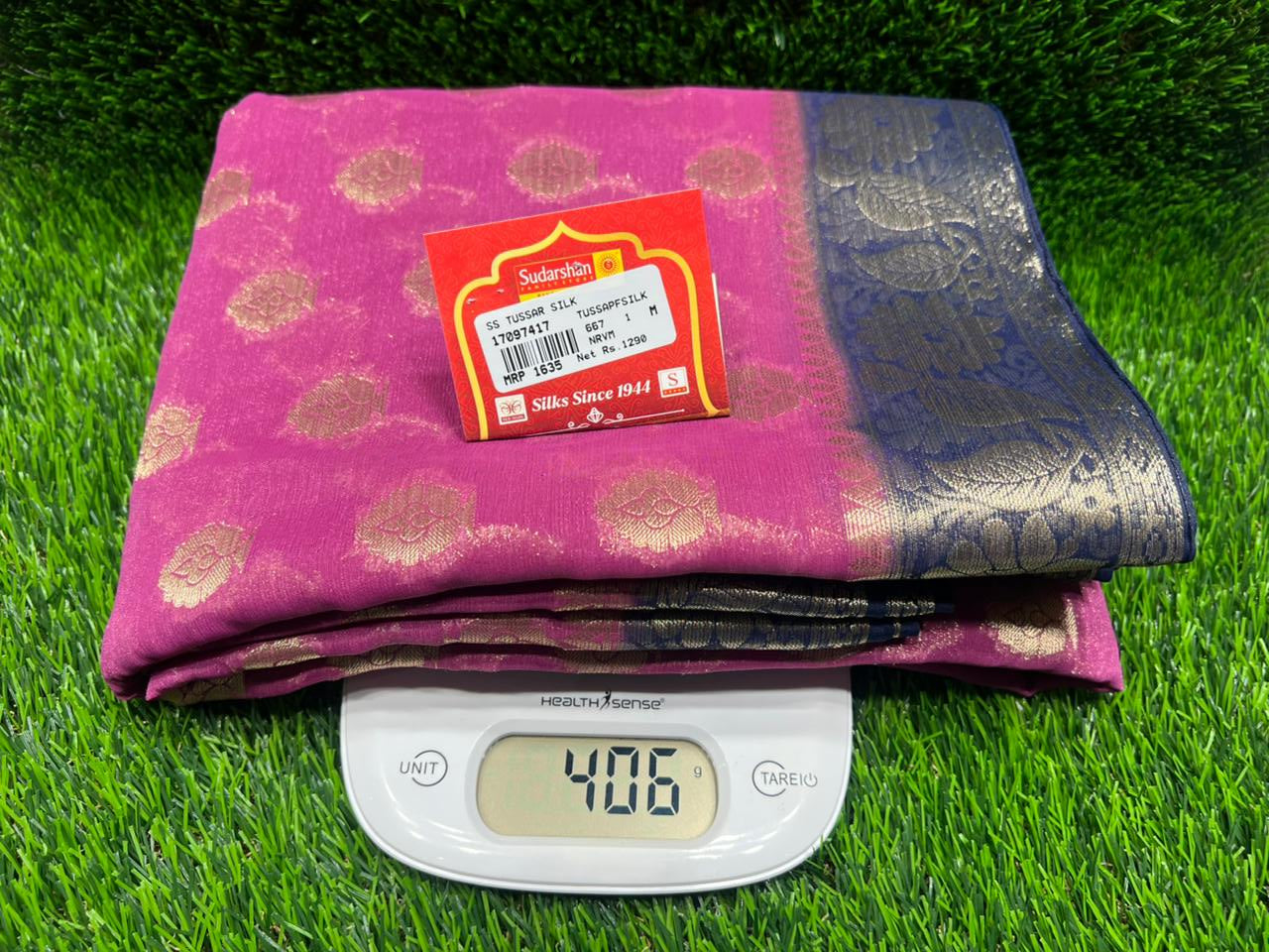 Pink fabric with a label on a green grass-like surface, weighed using a digital scale.