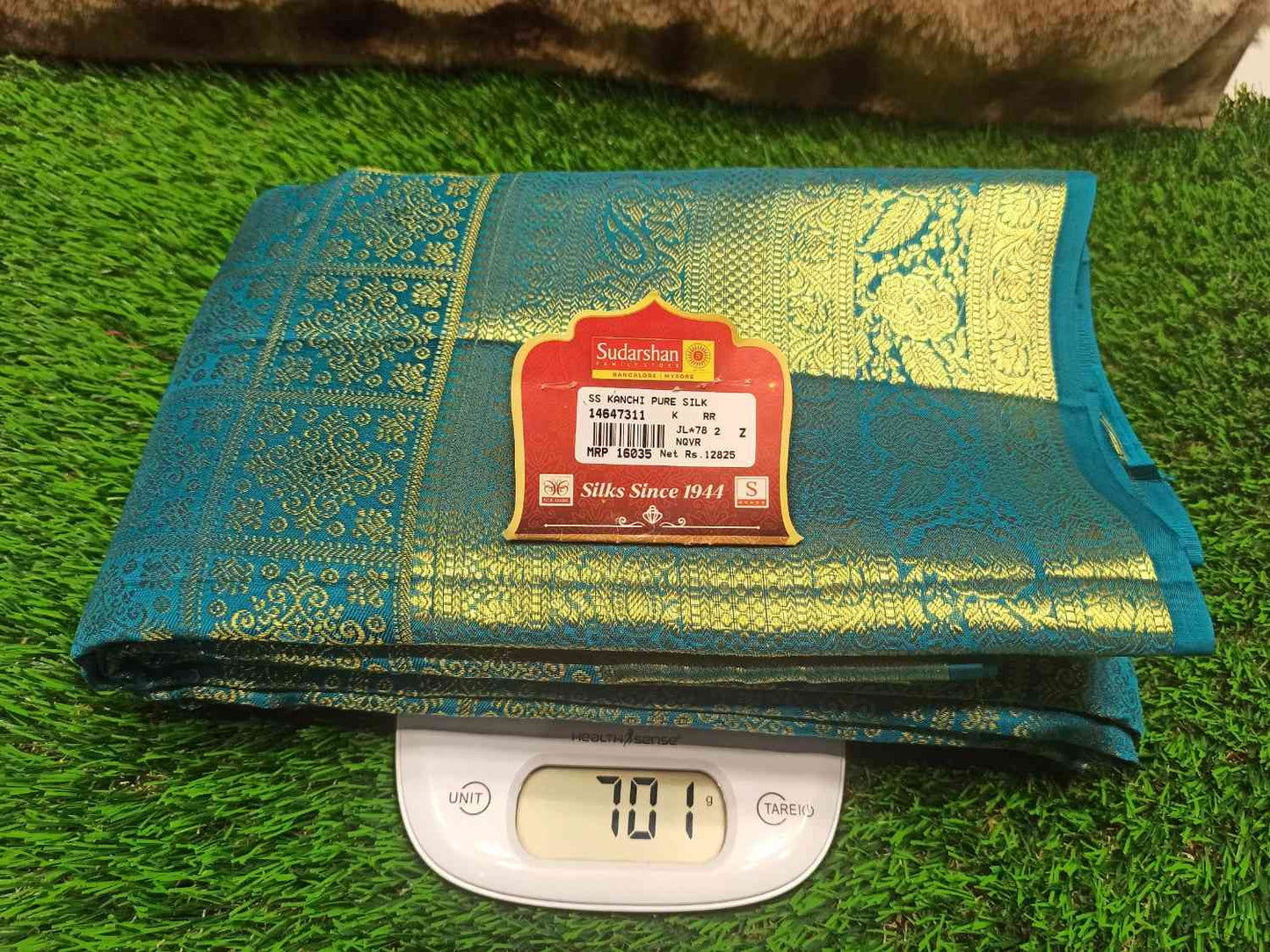 Folded green and gold fabric with a label on a digital scale against a grassy background
